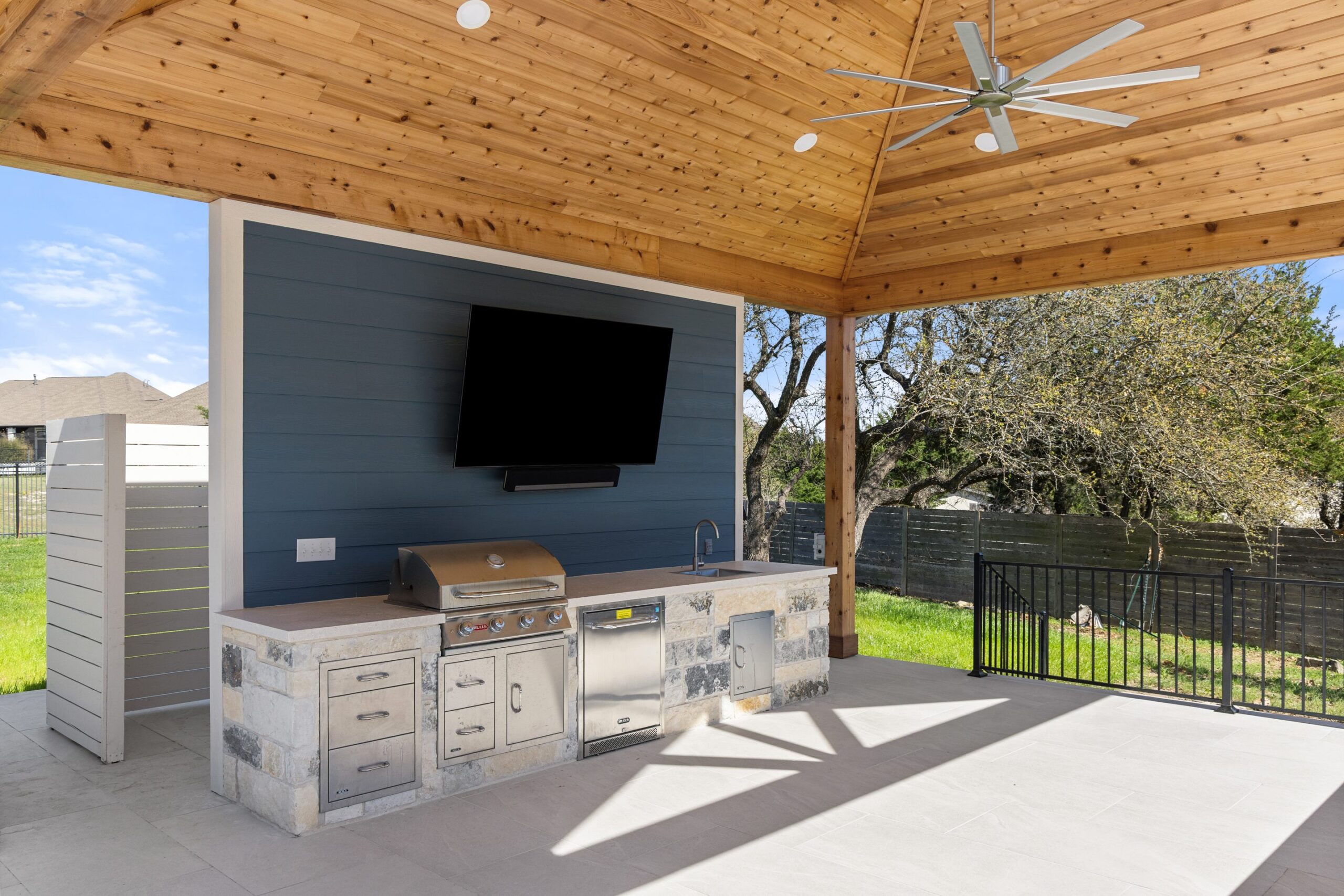 Interior view of Stratus Grove outdoor kitchen pavilion with stainless grill, mini fridge, TV wall, and ceiling fan, Wild Basin Pools Austin TX