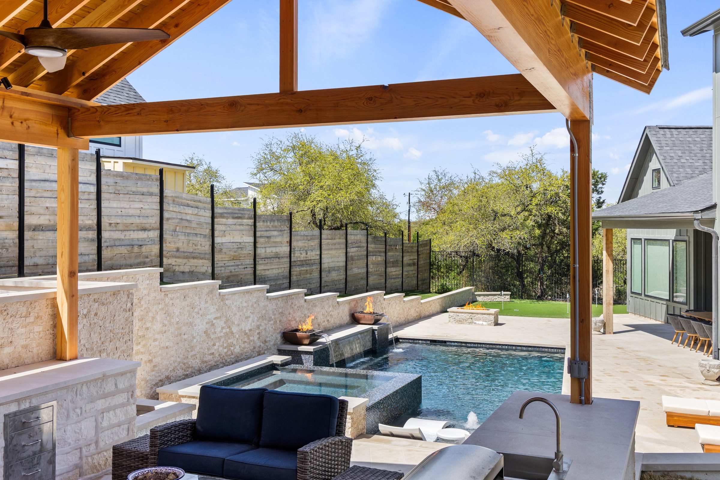 View from inside the cedar A-frame pavilion looking out toward The Commons pool and spa with copper fire bowls lit by Wild Basin Pools Austin TX